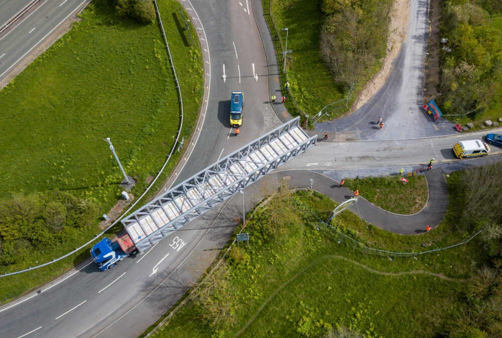 Aerial view of a Beaver Bridges truck transporting a massive steel truss section across a road junction, supported by an escort vehicle.