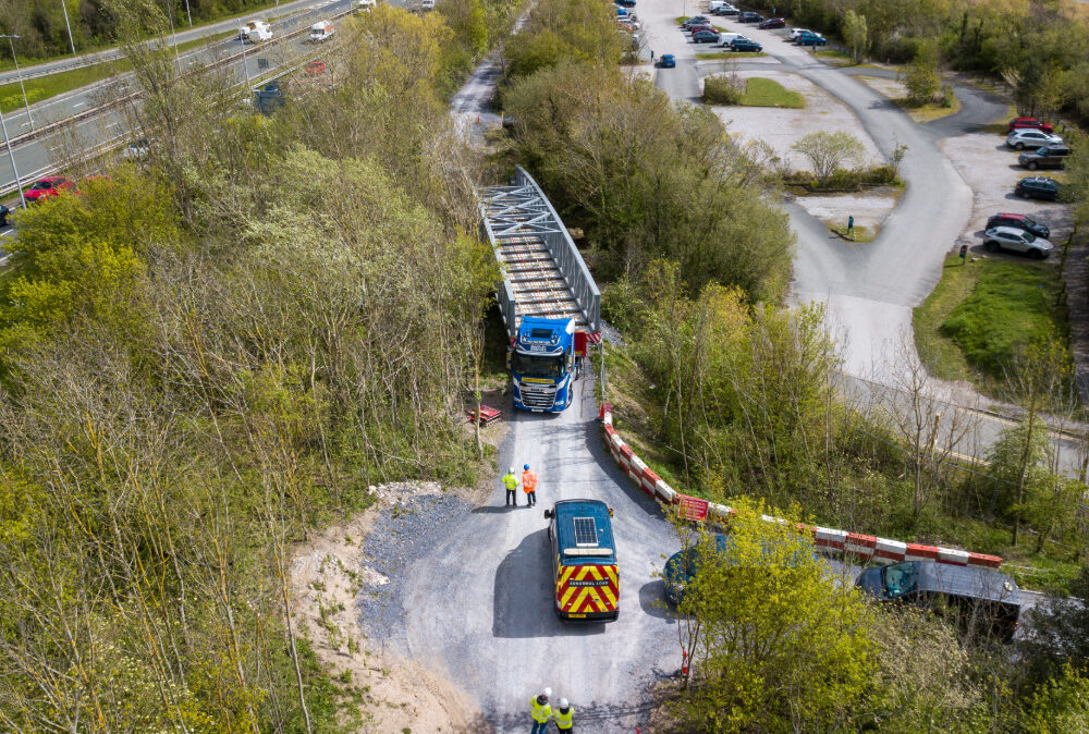 Aerial view of a Beaver Bridges truck carrying a steel truss bridge section on a narrow road, guided by an escort vehicle and ground crew.
