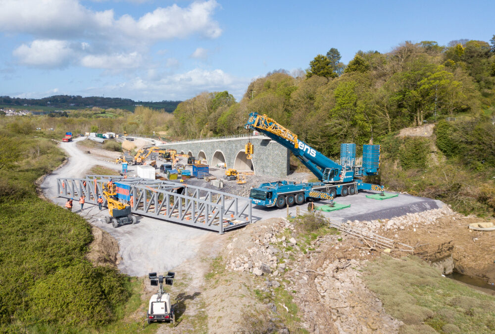 Aerial view of a Beaver Bridges site featuring a large Sarens crane, bridge truss sections, and a stone arch structure in a rural landscape.