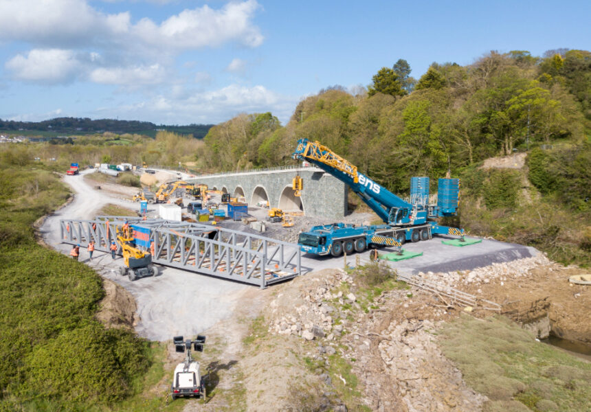 Aerial view of a Beaver Bridges site featuring a large Sarens crane, bridge truss sections, and a stone arch structure in a rural landscape.