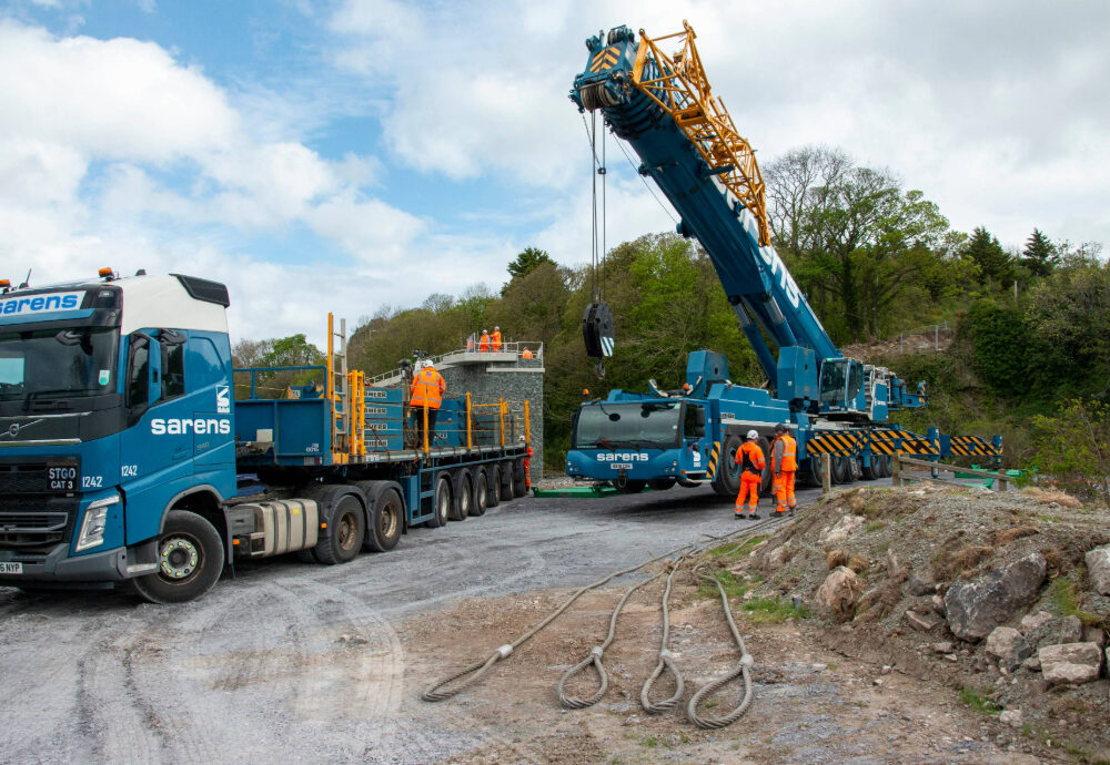 A blue Sarens crane and Volvo heavy-haulage truck on a gravel construction site, prepared for a Beaver Bridges installation project.