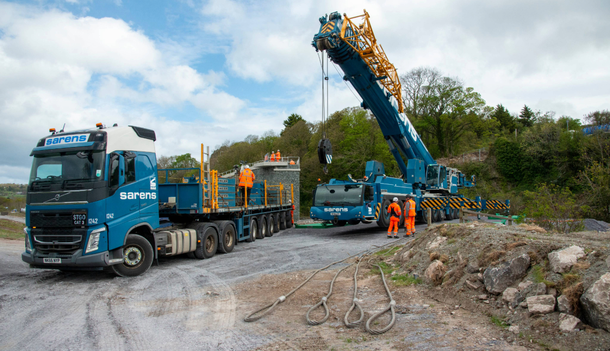 A blue Sarens mobile crane and Volvo transport truck on a Beaver Bridges site, with heavy lifting cables in the foreground and a stone bridge pier.