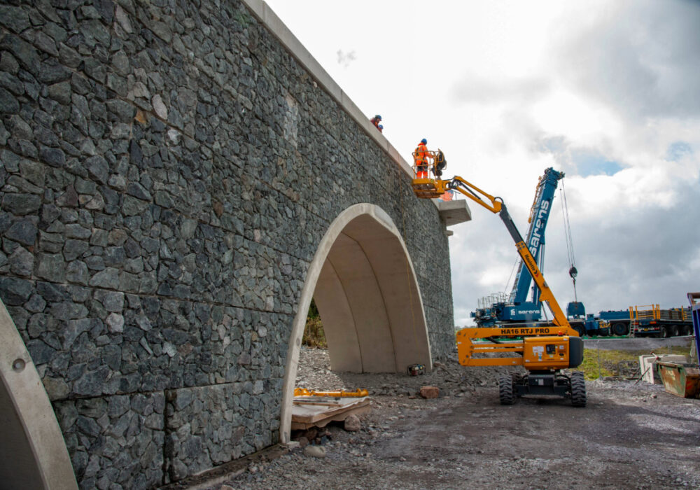 Workers on a cherry picker finalise a stone-clad arch bridge, with a large Sarens mobile crane and support equipment in the background