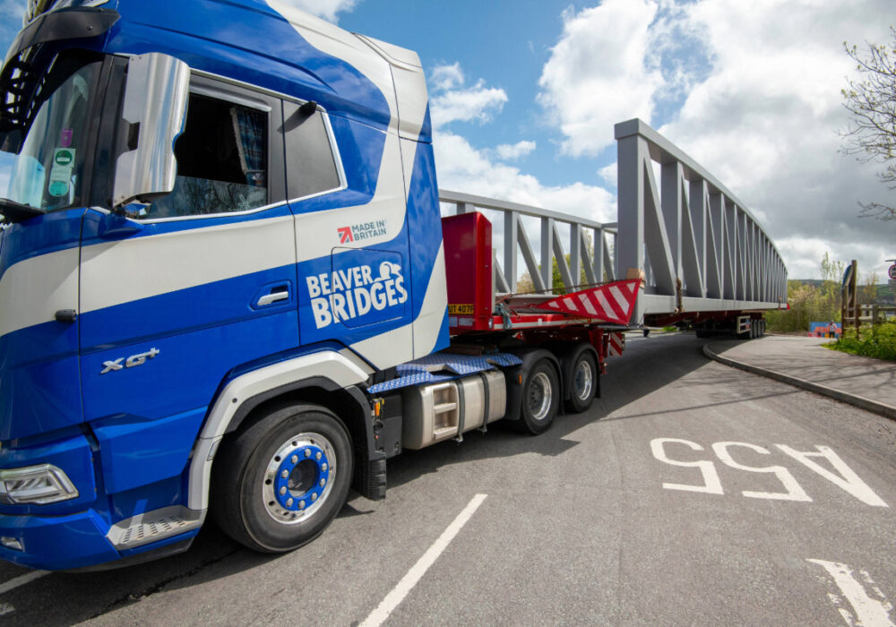 A blue and white branded Beaver Bridges truck carrying a large grey steel bridge section on a trailer, showcasing British manufacturing.
