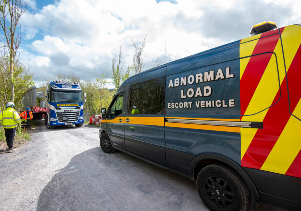 Beaver Bridges escort van and heavy-haulage truck transporting a bridge section on a narrow gravel road, with workers in hi-vis gear.