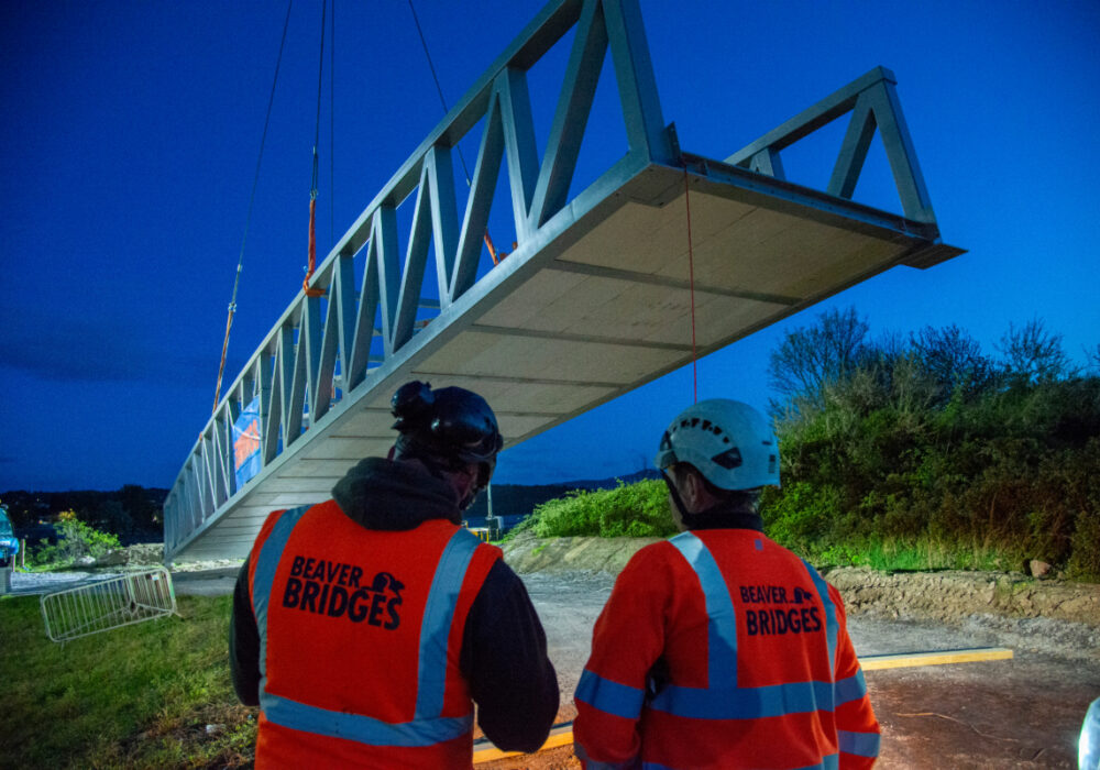 Two Beaver Bridges engineers in orange hi-vis vests watch as a large steel bridge section is lifted by crane during a night installation.