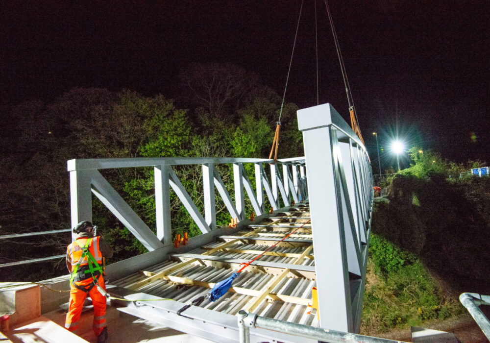 A Beaver Bridges engineer in safety harness monitors a steel truss bridge being lowered into place by crane during a nighttime installation.